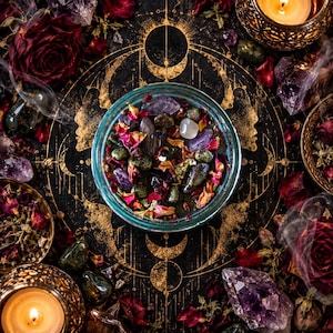 Overhead view of the Someone Fucked Up crystal intention candle centered over an elaborate gold lunar symbol on a dark textured altar cloth. The turquoise jar reveals dark wax covered with amethyst, bloodstone, dried rose petals, and assorted herbs. Surrounded by purple crystals, crimson flowers, and candlelight, this photograph showcases the candle's decorative toppings and mystical witchcraft aesthetic.