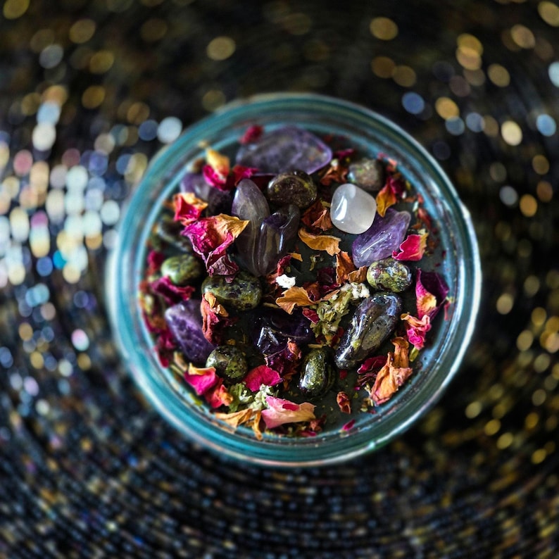 Overhead view of the Someone F'd Up candle showing a richly decorated wax top filled with dried rose petals, dark botanical herbs, colorful flowers, crystal chips, and small stones. Keywords: ritual candle, herbs and crystals, witchcraft candle, spellwork, intention candle.