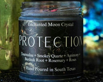 Peut inclure: Une bougie bleu foncé dans un pot en verre avec une étiquette noire portant le texte Enchanted Moon Crystal, PROTECTION, Tourmaline + Smokey Quartz + Agrimony + Burdock Root + Rosemary + Roses, et Hand Poured in South Texas avec des motifs floraux.
