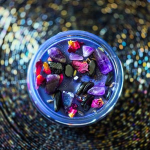 Overhead photograph of Witching Hour intention candle featuring deep purple wax, dark crystals, colorful rose petals and dried ritual herbs.