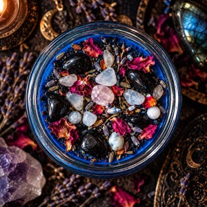 Overhead close-up of a handmade Hecate goddess candle showing brilliant deep blue wax decorated with glossy black obsidian stones, white and translucent moonstone crystals, vibrant dried rose petals, and scattered herbs. The candle is surrounded by shimmering blue labradorite, purple crystals, ornate golden celestial decorations, and lavender against a dark altar background. A richly embellished crystal candle inspired by Greek mythology, goddess worship, intuition, witchcraft, and moon magic.
