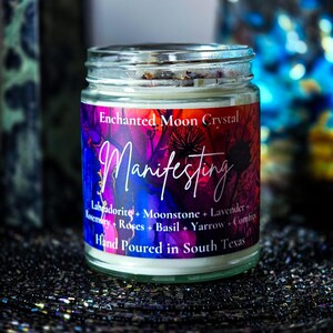 Op de afbeelding: Een glazen potkaars met een kleurrijk etiket. Het etiket luidt "Enchanted Moon Crystal Manifesting" en bevat ingredi&euml;nten zoals Labradoriet, Maansteen en Lavendel. De kaars is met de hand gegoten in Texas.