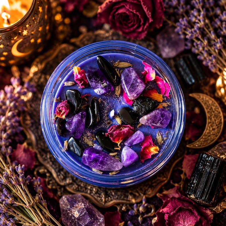 A round glass candle container filled with blue wax, topped with purple amethyst crystals, black obsidian stones, and dried pink rose petals. The candle surface is decorated with scattered botanical elements and polished gemstones.