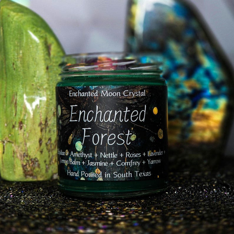 Może przedstawiać: Zielona świeca w szklanym słoiku z napisem "Enchanted Forest" na czarnej etykiecie. Etykieta zawiera r&oacute;wnież napis "Enchanted Moon Crystal" i listę składnik&oacute;w. Świeca jest ręcznie wylewana w południowym Teksasie.