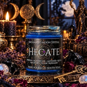 Hecate goddess intention candle by Enchanted Moon Crystal, featuring deep midnight blue wax in a glass jar with an ornate black and gold celestial label. Displayed on a mystical altar surrounded by glowing candles, crystals, dried flowers, and an antique gold triple moon symbol. This handcrafted witchcraft-inspired candle contains obsidian, moonstone, lavender, roses, mugwort, thyme, and rosemary, celebrating the Greek goddess of crossroads, protection, intuition, and transformation.