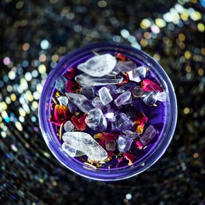 Pu&ograve; includere: Una candela viola in un contenitore di vetro trasparente, adornata con cristalli trasparenti e viola, petali di fiori rossi e gialli ed erbe. La candela poggia su una superficie scura e scintillante, creando un'estetica mistica.