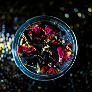 Top view of the handmade Banishing candle with dark wax, colorful dried botanicals, herbs and crystal accents arranged around the wick. Created for banishing work, spiritual cleansing, energy clearing, protection and releasing lingering unwanted energy.