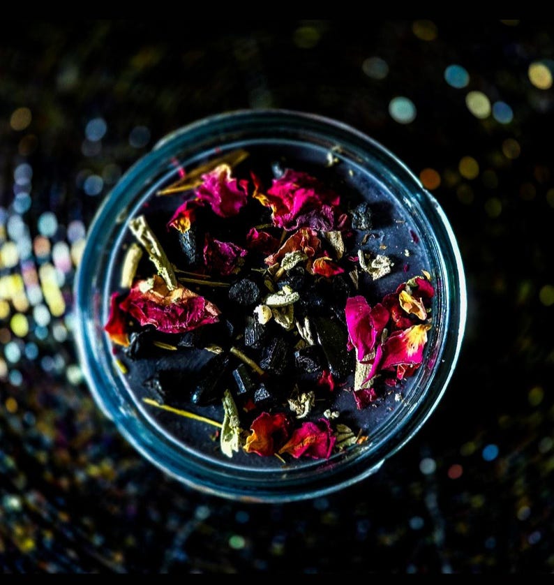 Top view of the handmade Banishing candle with dark wax, colorful dried botanicals, herbs and crystal accents arranged around the wick. Created for banishing work, spiritual cleansing, energy clearing, protection and releasing lingering unwanted energy.