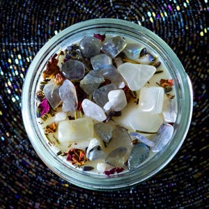 Op de afbeelding: Een glazen potkaars gevuld met witte was, versierd met diverse edelstenen en gedroogde bloemen. De kaars heeft een mix van grijze, witte en gele stenen, met vleugjes roze en paarse gedroogde bloemblaadjes. De achtergrond is een donker, getextureerd oppervlak.