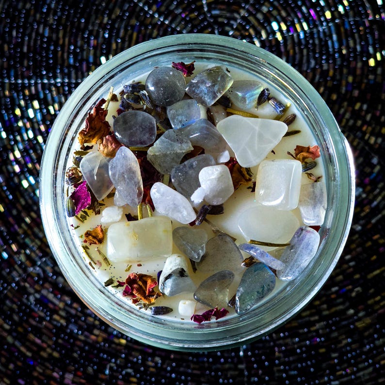 Op de afbeelding: Een glazen potkaars gevuld met witte was, versierd met diverse edelstenen en gedroogde bloemen. De kaars heeft een mix van grijze, witte en gele stenen, met vleugjes roze en paarse gedroogde bloemblaadjes. De achtergrond is een donker, getextureerd oppervlak.