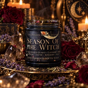 Direct front view of Season of the Witch handmade intention candle by Enchanted Moon Crystal, featuring a black glass jar with an original vintage gold moon label and dark wax topped with crystals and dried flowers. Displayed on an ornate antique gold pedestal surrounded by smoky quartz crystals, lavender, deep burgundy roses, and softly glowing candles. Luxurious gothic witchcraft-inspired product photography with rich amber lighting, celestial decor, and a mysterious magical atmosphere.