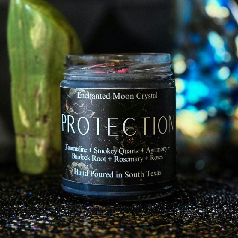May include: A black candle jar with a label that reads "Enchanted Moon Crystal PROTECTION Jourmaline + Smokey Quartz + Agrimony + Burdock Root + Rosemary + Roses Hand Poured in South Texas".