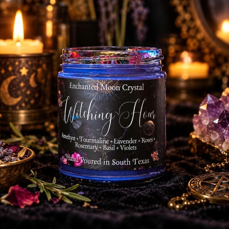 A blue glass candle jar with a black label featuring the text Enchanted Moon Crystal Witching Hour Amethyst + Tourmaline + Lavender + Roses + Rosemary + Basil + Violets Hand Poured in South Texas, topped with dried flowers.