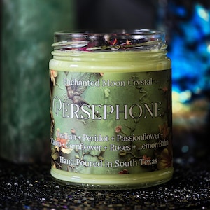 May include: A glass jar candle with a green label featuring the word "PERSEPHONE" and a hummingbird illustration. The label also lists ingredients like obsidian, peridot, and passionflower. The candle is hand-poured in South Texas.