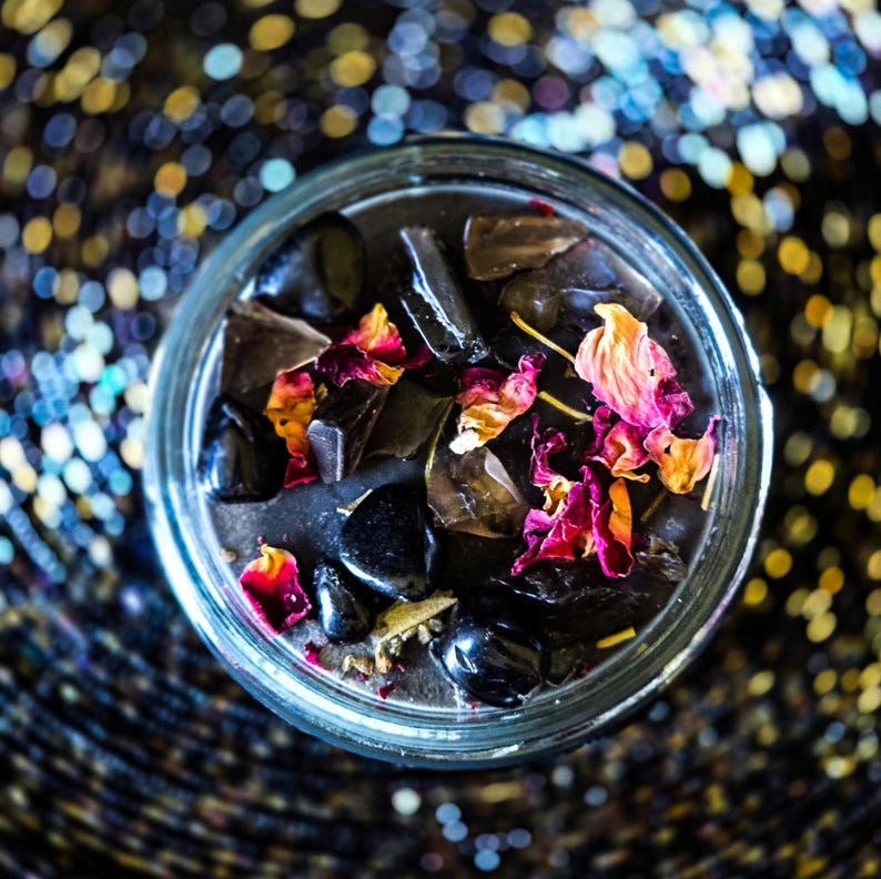 Close overhead photo of the Season of the Witch candle featuring glossy black wax decorated with smoky quartz, obsidian, dried rose petals and herbal botanicals. Dark witch candle handcrafted for ritual, spellwork, protection, grounding and spiritual practice.