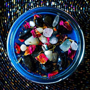 Overhead view of the Hecate intention candle with deep blue wax topped with Moonstone, Lavender, Mugwort, Thyme, and Rosemary. Pale stones, red and pink botanical accents, and natural herbs create a striking magical design.
