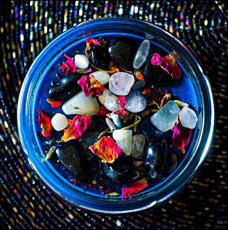 Overhead view of the Hecate intention candle with deep blue wax topped with Moonstone, Lavender, Mugwort, Thyme, and Rosemary. Pale stones, red and pink botanical accents, and natural herbs create a striking magical design.