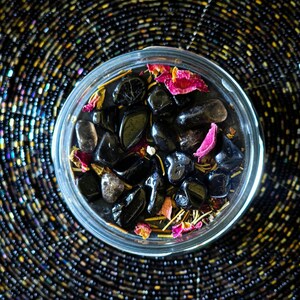 May include: A glass jar filled with black stones, dried rose petals, and other natural elements. The jar is sitting on a black and gold patterned surface.