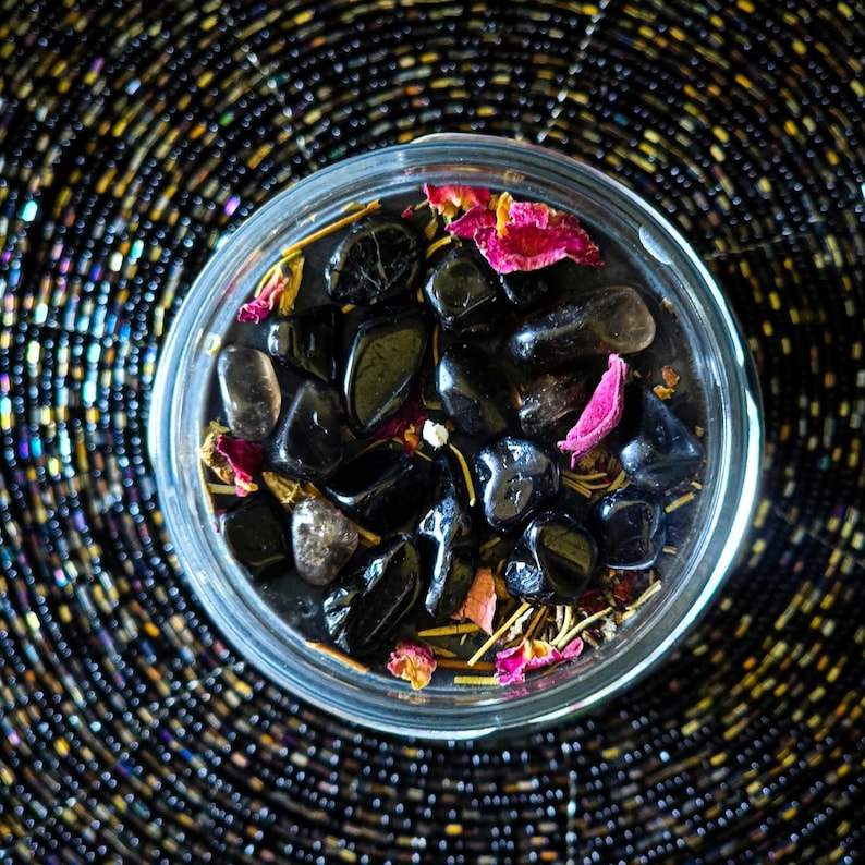 May include: A glass jar filled with black stones, dried rose petals, and other natural elements. The jar is sitting on a black and gold patterned surface.