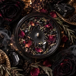 Enchanted Moon Crystal Banishing candle featuring black wax decorated with dried pink and burgundy rose petals, black tourmaline stones and a botanical blend of rosemary, sage, wormwood, horehound and hyssop. Displayed among black roses, dark crystals, scattered herbs and atmospheric ritual smoke. This handcrafted gothic witchcraft candle is designed for symbolic banishing, spiritual protection, cleansing negative energy, shadow work and intention setting. Dark mystical altar aesthetic