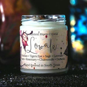 May include: A clear glass candle jar with a white wax fill and a label featuring the text Enchanted Moon Crystal, Lowak, Obsidian, Tigers Eye, Sage, Lavender, Roses, Rosemary, Chamomile, Damiana, and Hand Poured in South Texas.