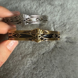 May include: Two metal cuff bracelets featuring a central fan-shaped motif and geometric diamond patterns. One bracelet is finished in a polished silver tone, while the other is a gold tone with black enamel detailing along the band and side accents.