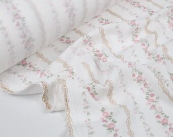 May include: White fabric roll with a floral pattern. The fabric features vertical stripes of pink roses and green leaves, interspersed with tan decorative borders. The fabric is soft and flowing, ideal for sewing projects.