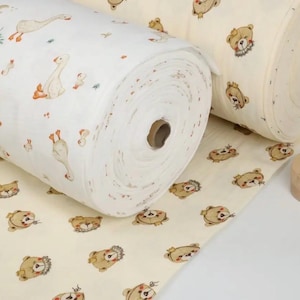 May include: Two rolls of fabric with whimsical patterns. One roll features geese and small floral accents on a white background. The other roll displays teddy bears on a cream background. A wooden stamp is also visible.