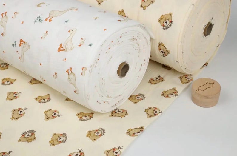 May include: Two rolls of fabric with whimsical patterns. One roll features geese and small floral accents on a white background. The other roll displays teddy bears on a cream background. A wooden stamp is also visible.
