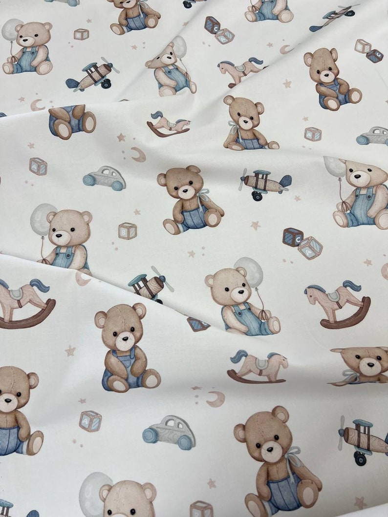 Pu&ograve; includere: Tessuto bianco con un motivo ripetuto di orsacchiotti con salopette blu, aeroplani, cavalli a dondolo, macchinine e blocchi alfabetici. Gli orsi tengono in mano dei palloncini. Il disegno comprende stelle e mezzelune.