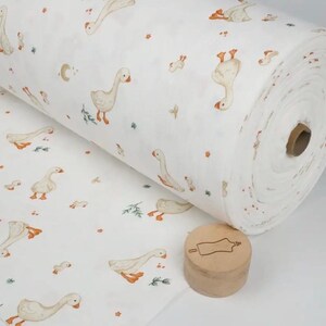 May include: A roll of white fabric featuring a repeating pattern of geese in various poses, with orange beaks and feet. The fabric also has small floral and leaf accents. A wooden stamp is in the foreground.