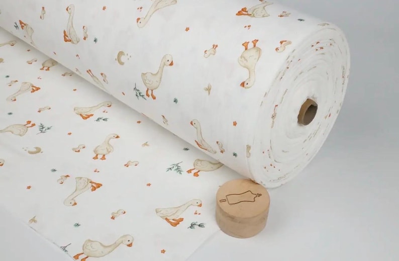 May include: A roll of white fabric featuring a repeating pattern of geese in various poses, with orange beaks and feet. The fabric also has small floral and leaf accents. A wooden stamp is in the foreground.