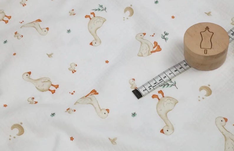 May include: White fabric with a repeating pattern of geese, ducklings, and botanical elements in shades of beige, orange, and green. A wooden spool with a dress form icon and a measuring tape are also visible.