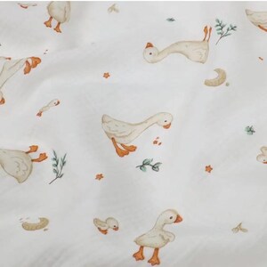 May include: White fabric with a repeating pattern of geese in various poses. The geese are depicted in shades of beige, with orange feet and beaks. Small green sprigs and orange star shapes are scattered across the fabric.