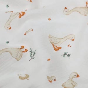 May include: White fabric with a repeating pattern of watercolor geese in various poses. The geese have orange beaks and feet. Scattered among the geese are small orange stars, green sprigs, and crescent shapes.
