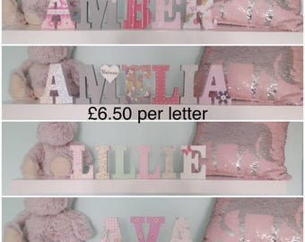May include: A selection of decorative wooden letters spelling out names, in various colors and patterns. The letters are displayed on a white shelf, with a teddy bear and a sequined pillow in the background. The names include "AMBER", "AMELIA", "LILLIE", and "AVA".
