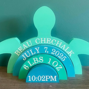 Peut inclure: Un souvenir en bois en forme de tortue, de couleur bleu sarcelle et bleu, personnalisé avec le nom "Beau Chechalk", la date de naissance du 7 juillet 2025, le poids de 2,72 kg et l'heure 22h02. Le design présente des arches superposées.