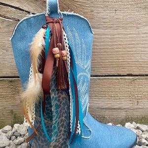 May include: A light blue denim cowboy boot featuring white stitched patterns and a decorative charm accessory attached to the pull tab. The accessory includes brown ribbons, turquoise satin strips, white lace, feathers, and wooden beads.