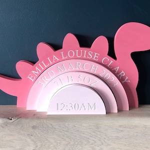 May include: Pink wooden dinosaur shaped baby birth announcement with the text "Emilia Louise Clary, 3rd March 2023, 7lb 5oz, 12:30AM"