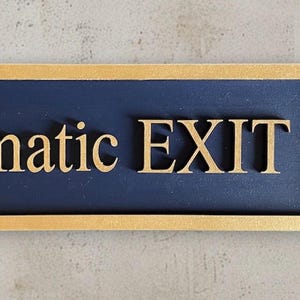 May include: A rectangular sign with a gold frame and the words "Dramatic EXIT" in gold lettering. The background is navy blue. The sign is mounted on a light gray wall, with a plant visible on the right side.
