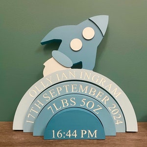 May include: A blue and white wooden rocket ship with a rainbow-colored archway with the text "Olly Ian Ingram 17th September 2024 7 LBS 5 OZ 16:44 PM"
