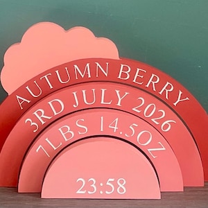 May include: A multi-layered wooden rainbow plaque in shades of red and pink with a cloud shape. The text reads AUTUMN BERRY, 3RD JULY 2026, 7LBS 14.5OZ, and 23:58 in white lettering. The object features four stacked semi-circular arches.