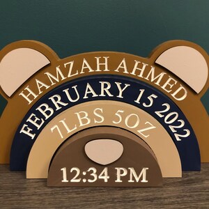 May include: A multi-layered wooden bear-shaped plaque featuring the name HAMZAH AHMED, the date FEBRUARY 15 2022, the weight 7LBS 5OZ, and the time 12:34 PM. The design uses shades of brown, tan, and navy blue with cream-coloured engraved text.