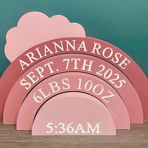 May include: A multi-layered wooden rainbow plaque in shades of pink featuring a cloud shape at the top. The text reads ARIANNA ROSE, SEPT. 7TH 2025, 6LBS 10OZ, and 5:36AM in white lettering across the four arched segments of the rainbow structure.
