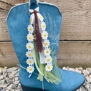 Könnte beinhalten: Ein blauer Jeansstiefel mit einem dekorativen Anhänger an der oberen Zugschlaufe. Der Anhänger besteht aus weißer Gänseblümchen-Spitze, braunen Wildlederquasten, Holzperlen und hellgrünen Bandstreifen, die an einem silbernen Metallring hängen.