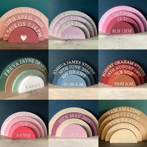 Puede incluir: Un conjunto de diez arcos de madera de colores arcoíris con texto personalizado, que incluye nombres, fechas, pesos y horas. Los arcos están apilados en un tamaño graduado, con el arco más grande en la parte inferior y el arco más pequeño en la parte superior. El texto es blanco y es visible contra el fondo de color.