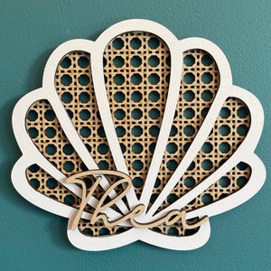 May include: White wooden seashell wall decor with a woven pattern and the word "Thea" written in cursive.