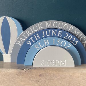Puede incluir: Decoración de madera con forma de arcoíris y globo aerostático. El arcoíris es azul, gris y blanco, con el nombre "PATRICK MCCORMICK", fecha de nacimiento, peso (3,6 kg) y hora (20:05) grabados.