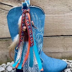 May include: A blue denim cowboy boot featuring a decorative charm attached to the pull tab. The charm consists of a collection of colorful ribbons, white lace, brown wooden beads, and feathers, creating a layered and textured accessory.