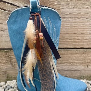 May include: A blue denim cowboy boot featuring a frayed top edge and white decorative stitching. A decorative charm hangs from the pull tab, consisting of brown and white feathers, a brown tassel, wooden beads, and leopard print ribbon.